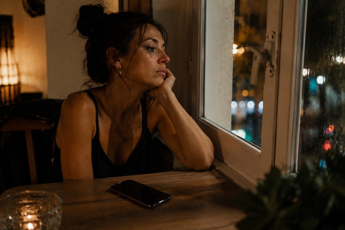 mujer sentada sola junto a una ventana después de una reunión social con maquillaje ligeramente desgastado y el celular apagado sobre la mesa