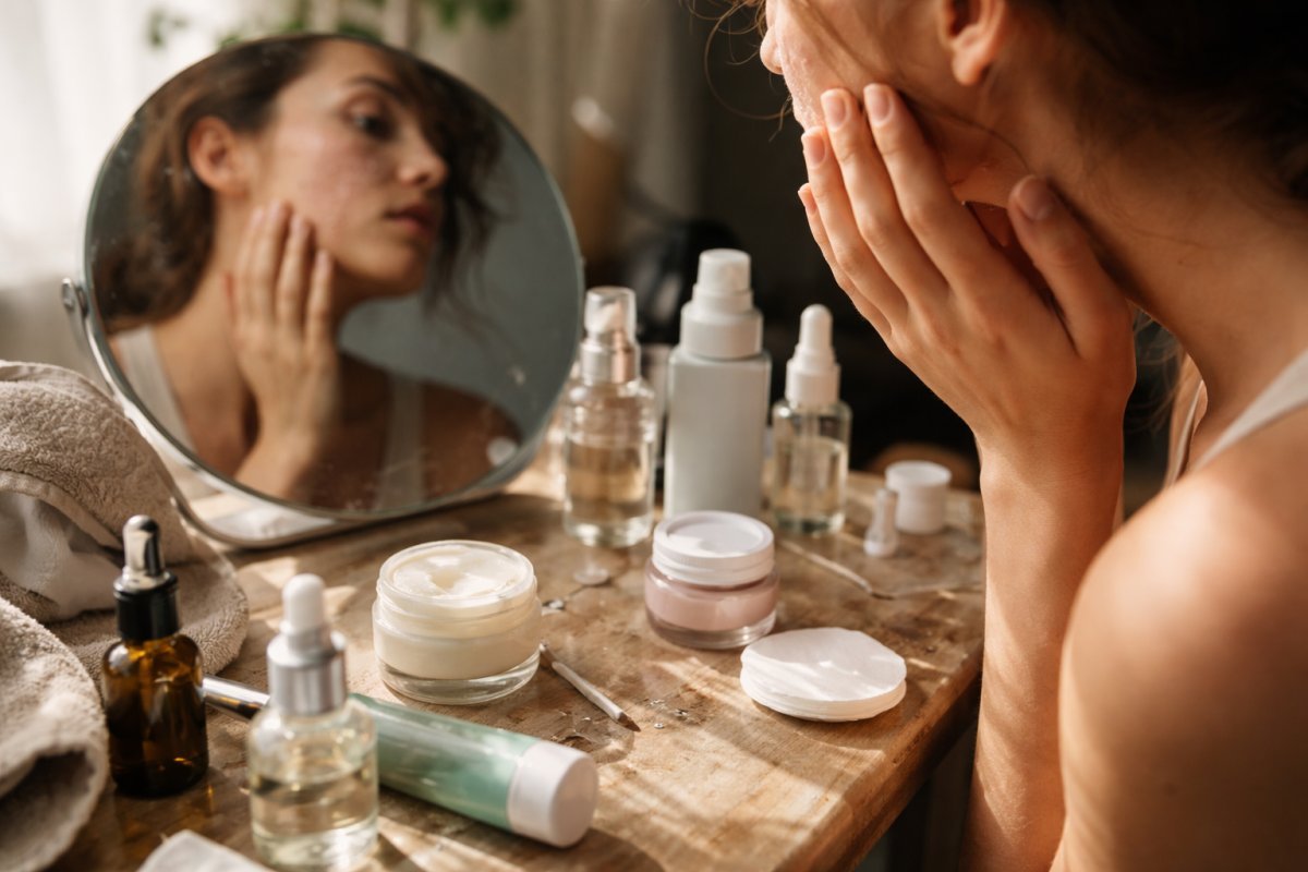 Frascos de skincare abiertos sobre una mesa desordenada junto a una piel con textura irregular frente al espejo